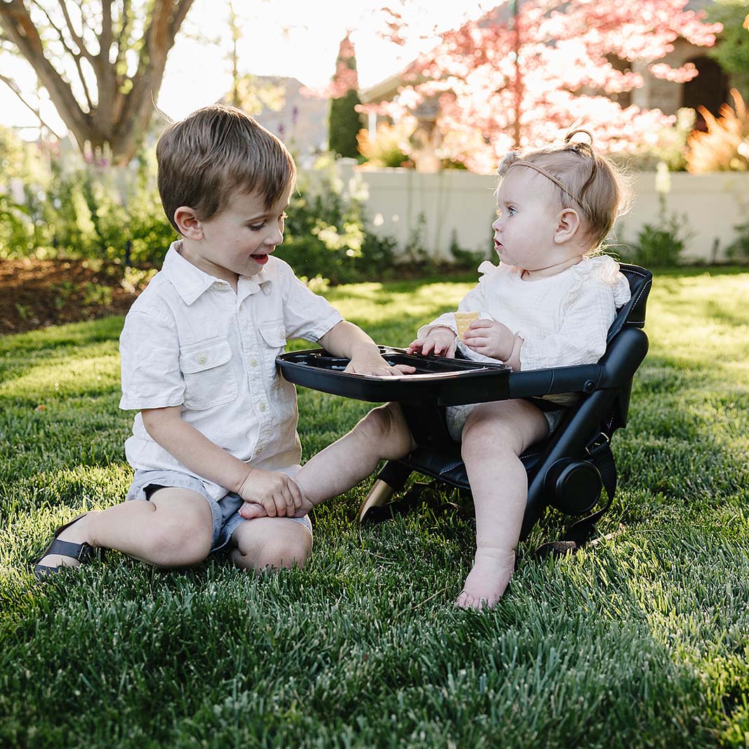 Two children playing together on a grassy area with a baby chair