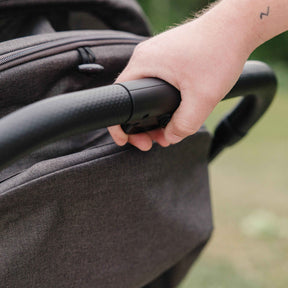Hand holding onto a stroller handle demonstrates the auto fold mechanism with a blurred natural background