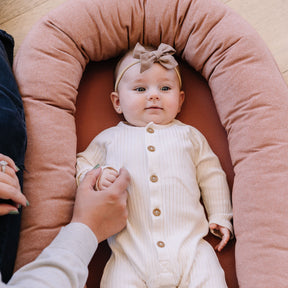 Baby in a white outfit with a bow sitting on a pink cushion, holding a hand.
