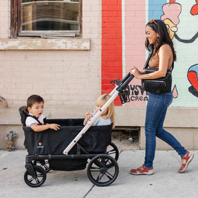 Woman pushing a black stroller wagon with two children in an urban setting.