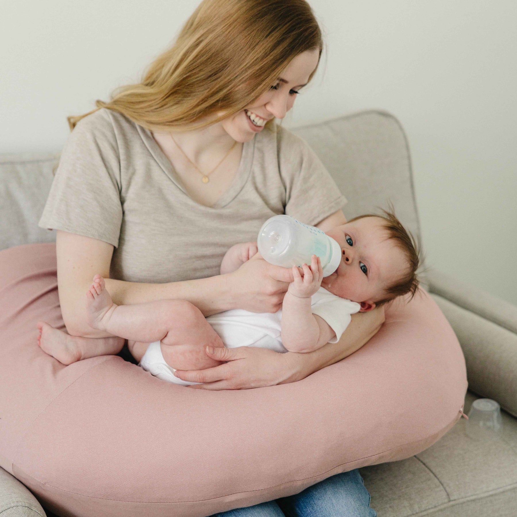 Woman holding a baby on a pink cushion, with a bottle in hand, in a cozy indoor setting.