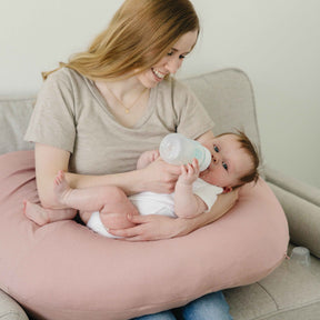 Woman holding a baby on a pink cushion, with a bottle in hand, in a cozy indoor setting.
