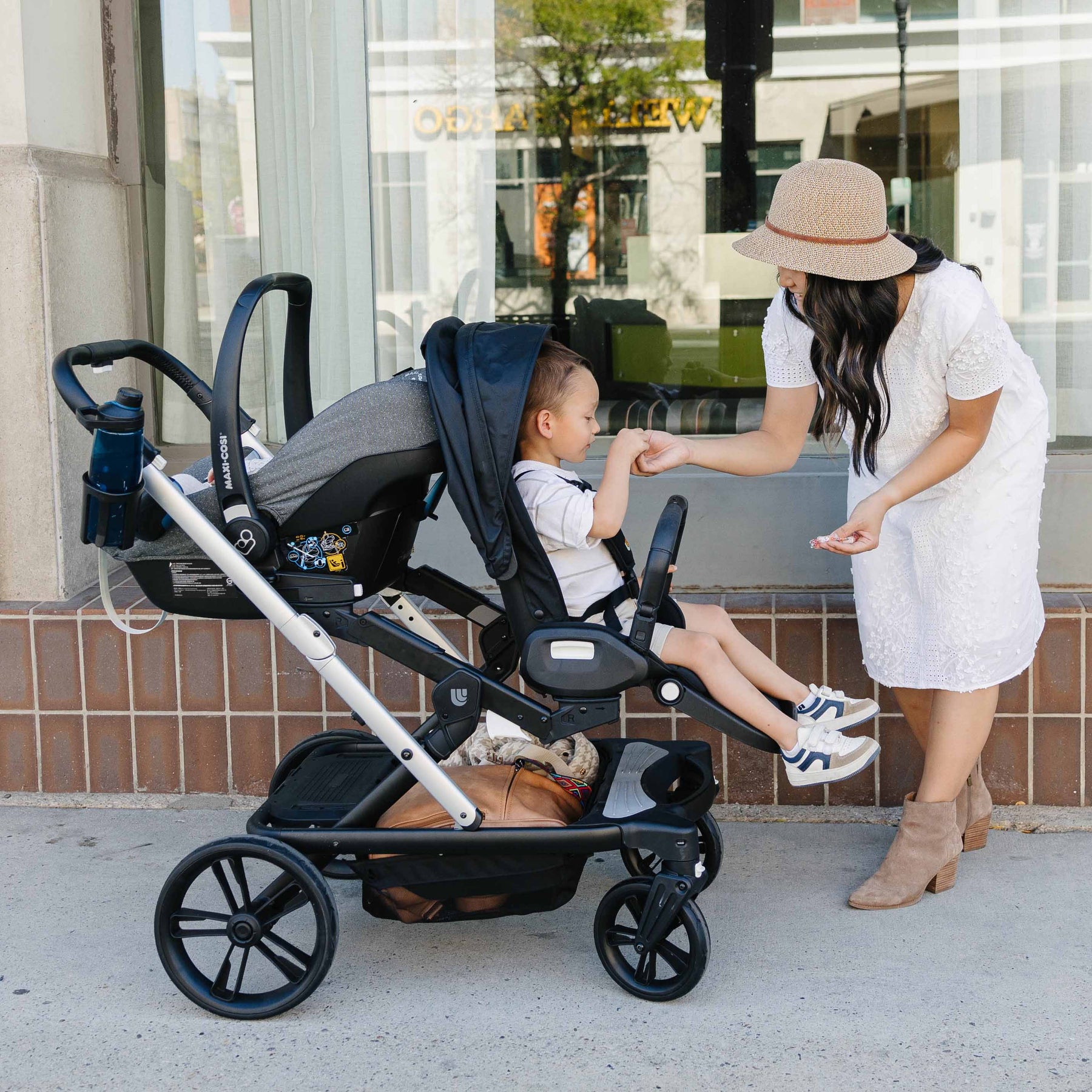 Woman in a white dress and hat interacting with a child in a stroller on a sidewalk.
