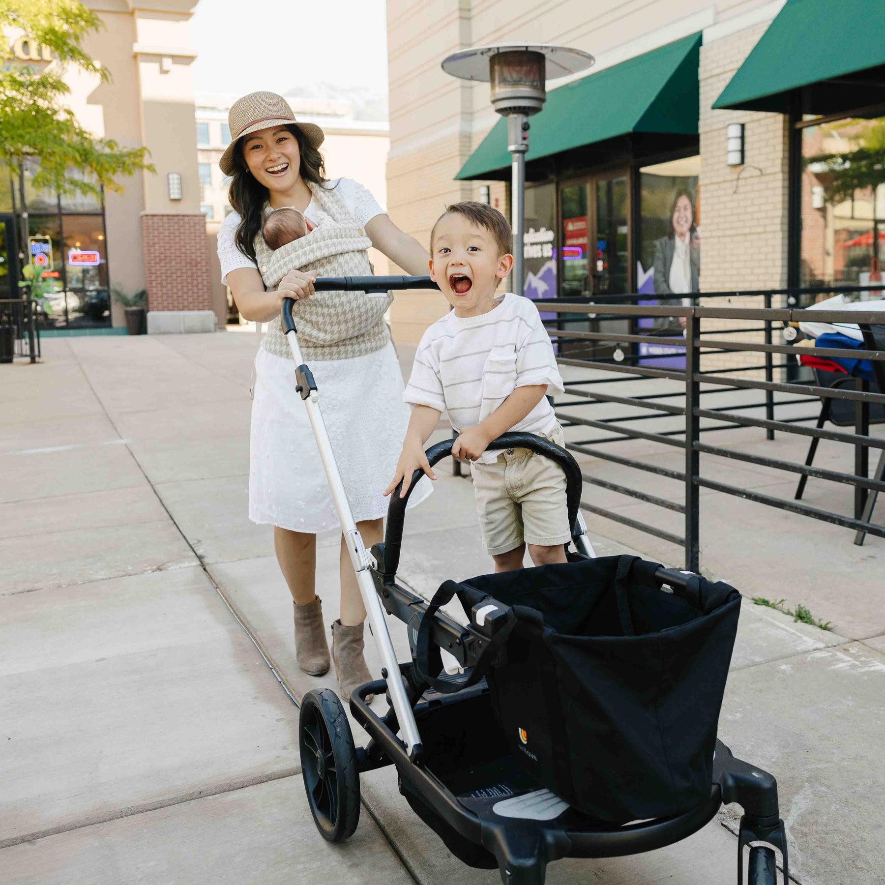 Woman pushing a stroller with a baby in a sling and a child standing next to it on a sidewalk.